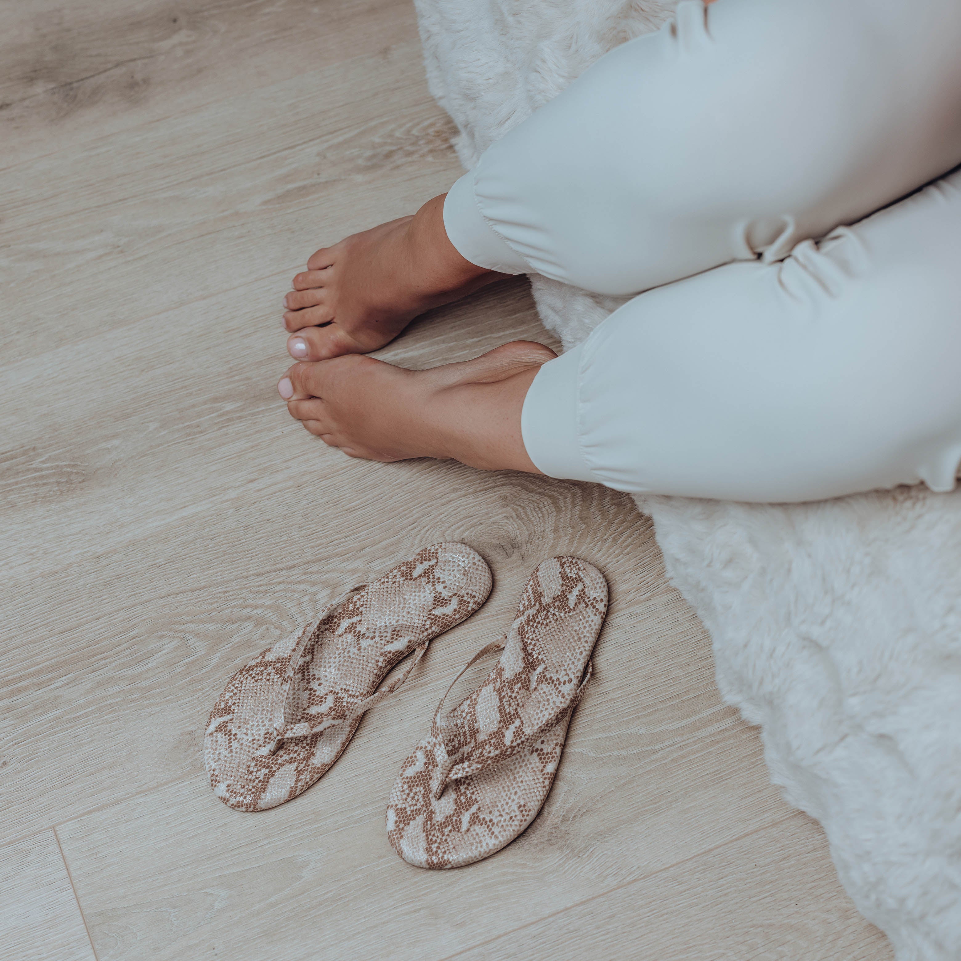 Snake print flip-flops on a wooden floor with a person wearing white pants.