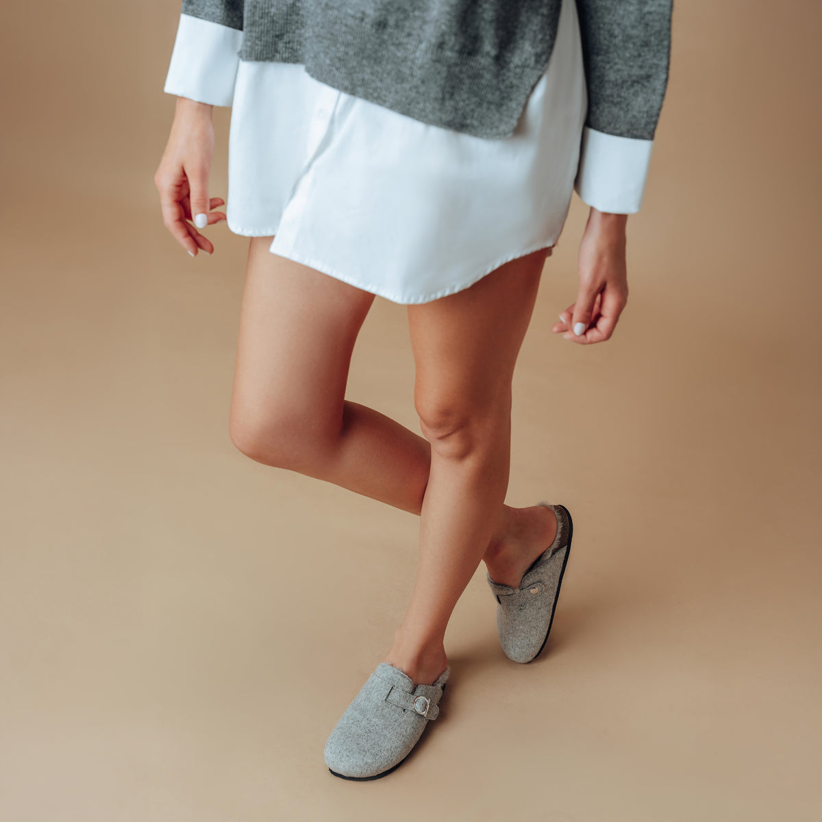 A person wearing gray knit slip-on clogs with a buckle strap, paired with a white mini skirt and gray sweater, standing against a solid beige studio background.