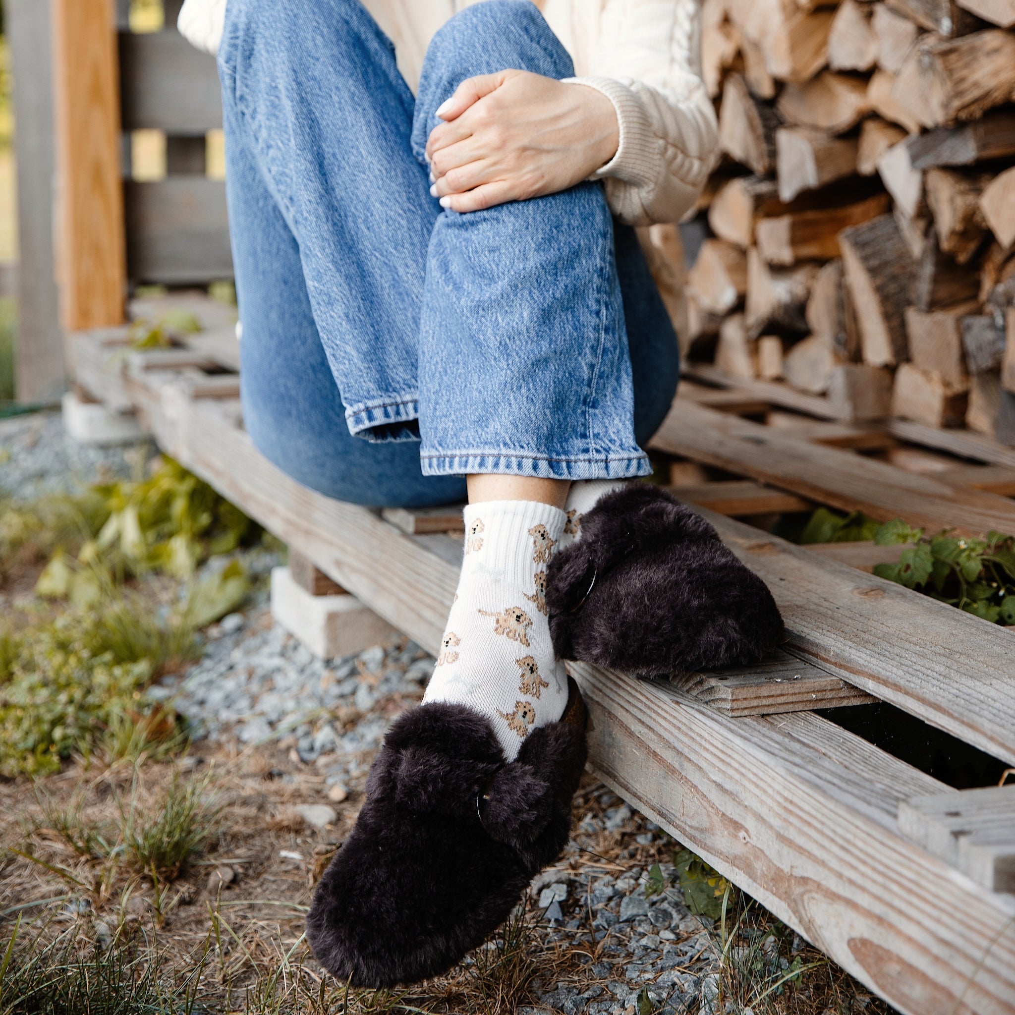 Woman sitting on a wooden bench wearing a white coat, blue jeans, and Aspen Bark Faux Fur Clogs by Solei.Sea